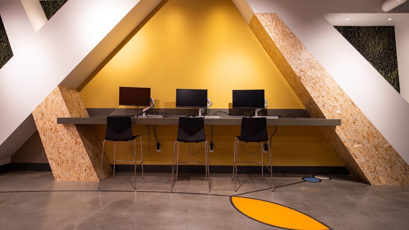An image of a row of computers on a counter with tall stools where people can work on the computers.