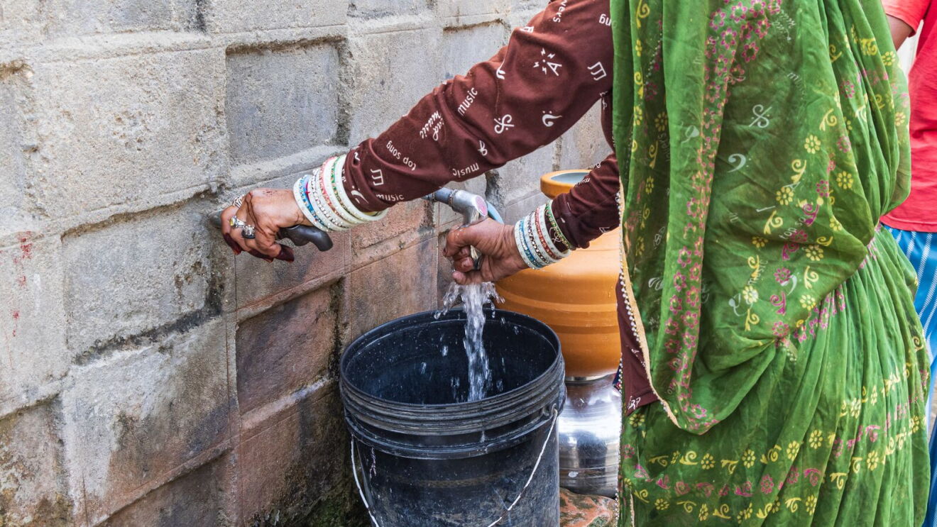 Woman in green sari and brown sleeve filling a black bucket from a wall-mounted water tap