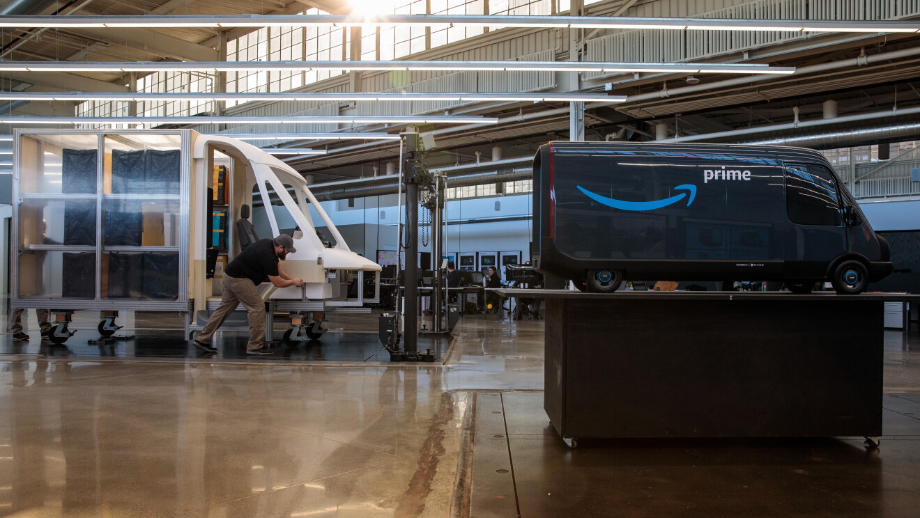 Modello in scala di un furgone Amazon, esposto su un tavolo. Sullo sfondo, lo scheletro del prototipo a grandezza naturale con un uomo. 