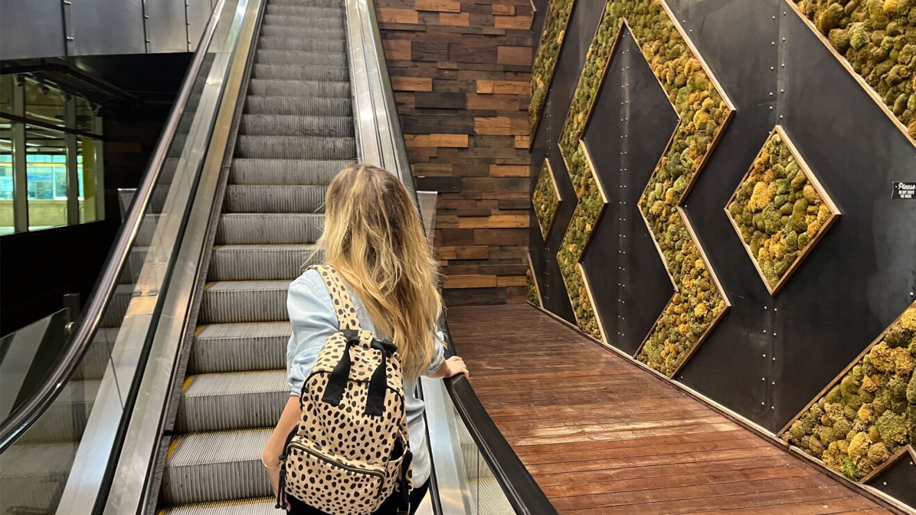 an image of a woman going up the stairs of an escalator at a Whole Foods store. There is a green and black living wall to her right and unique wood design panels along the rest of the walls. She is looking away from the camera and carrying a leopard print backpack.