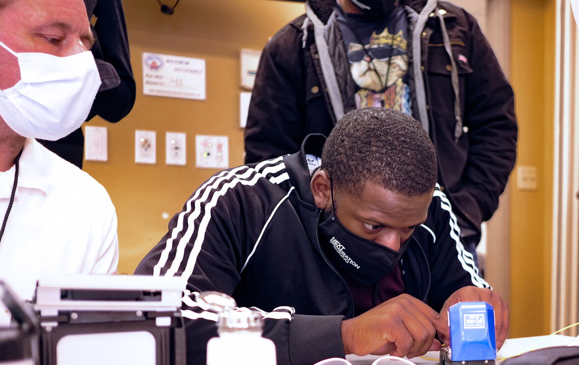 A man is sitting at a table working on a piece of technology hardware. His instructor and fellow students look on as he works. 