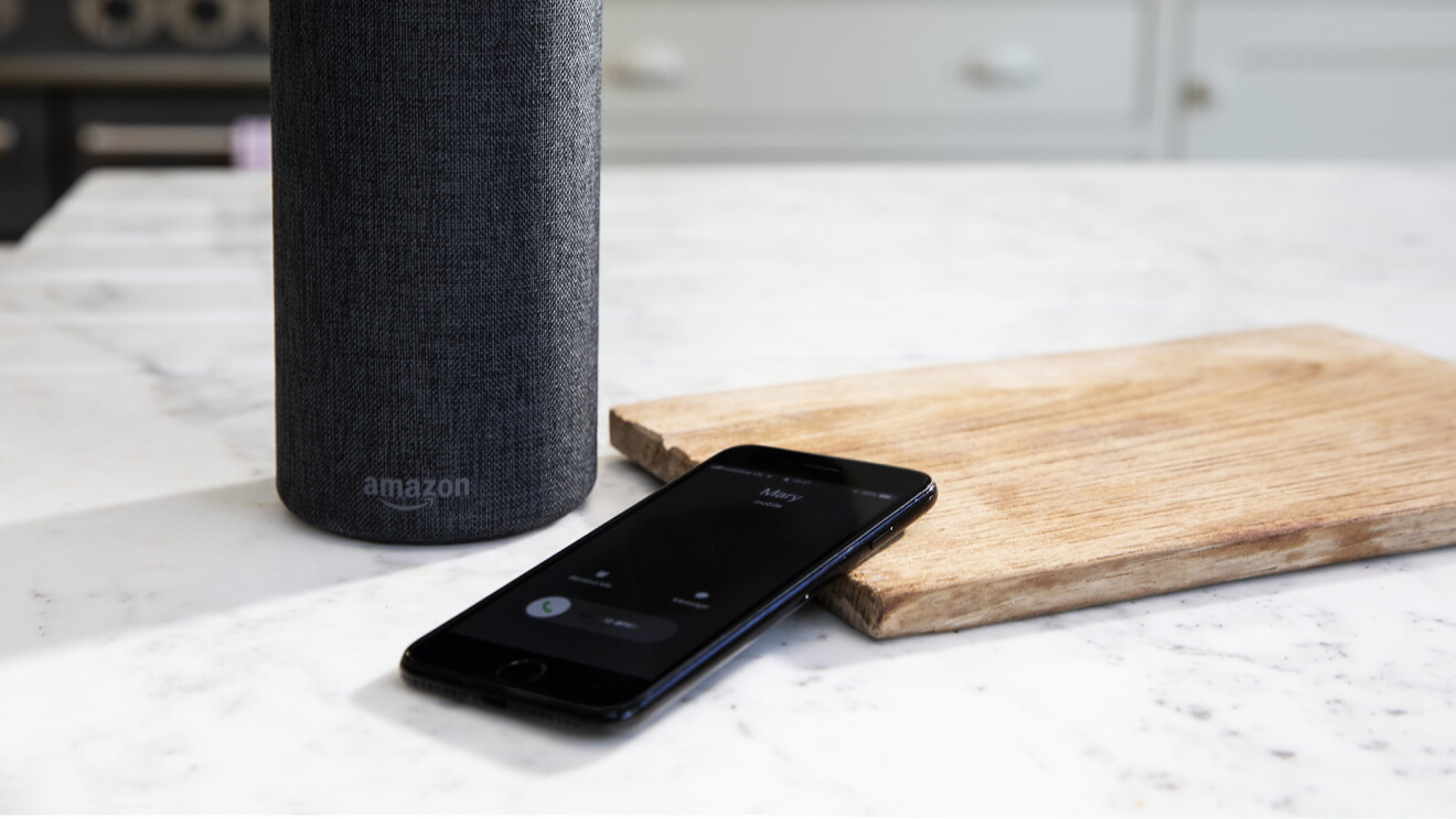 An Amazon Echo sits on a kitchen counter, in front of it, a cellular phone rests against a cutting board.