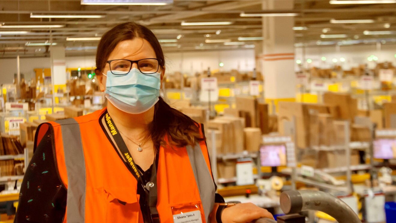 Eine Frau mit Brille, Mundschutz und braunen langen Haaren steht in einer Logistikhalle.