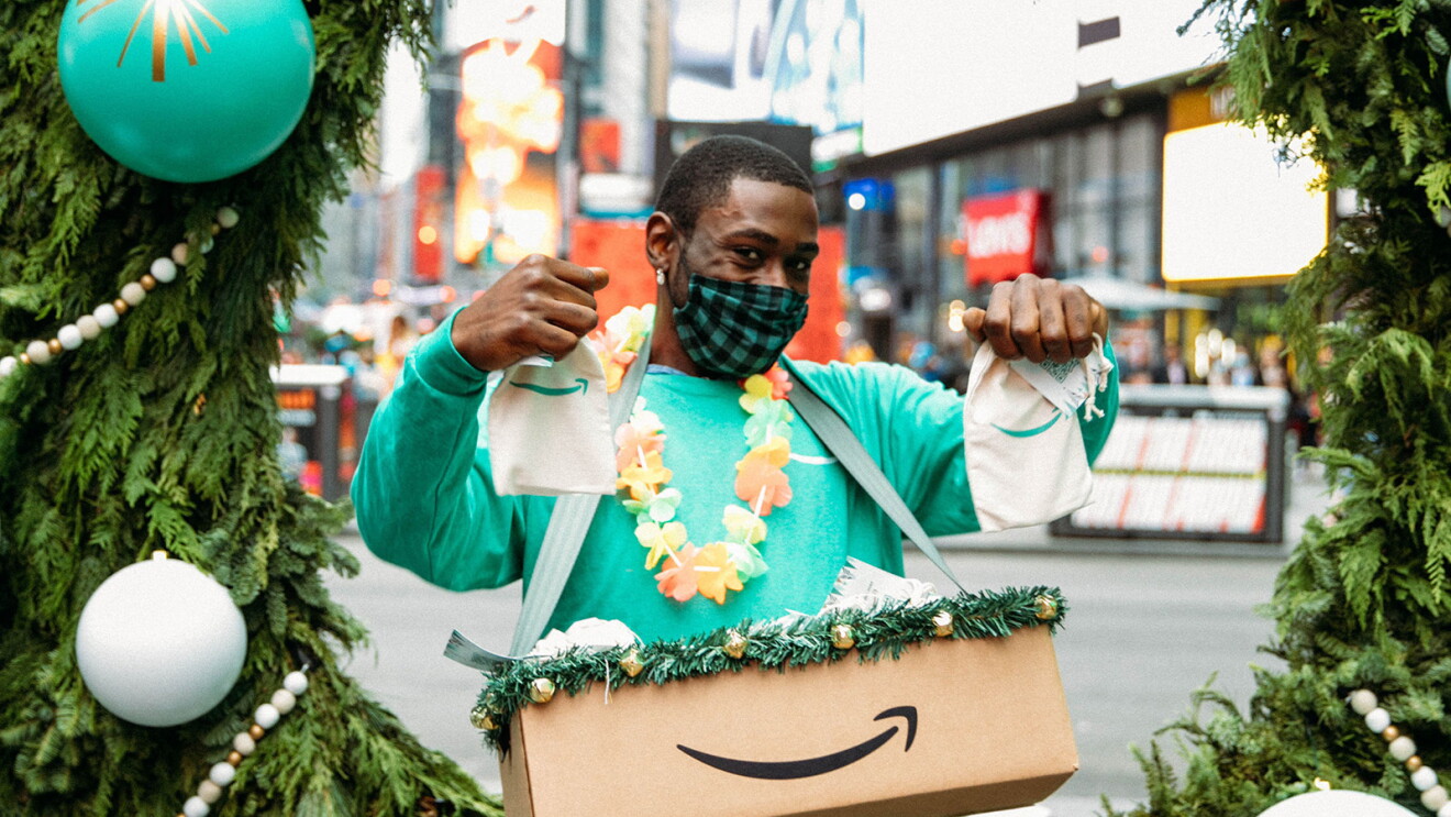 An image of a man smiling for a photo while wearing a mask. He is holding up two gift bags with the Amazon logo on them and has a box with the Amazon logo in front of him. 