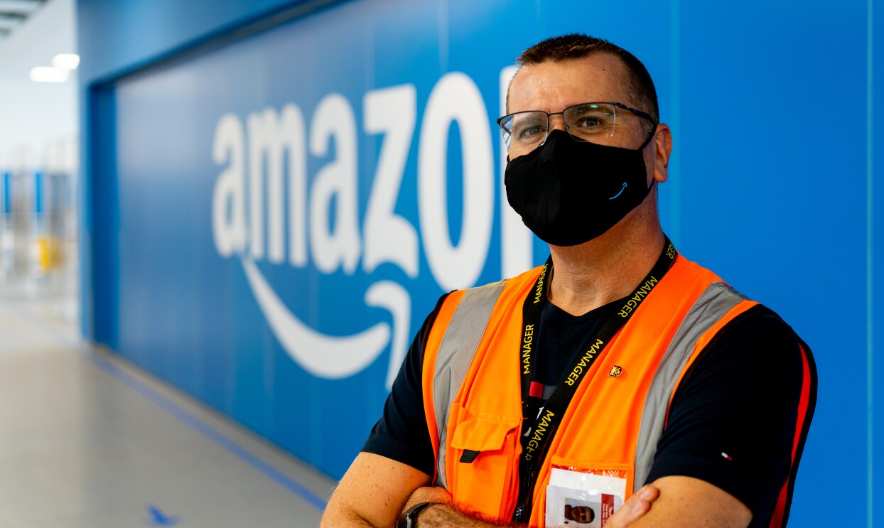 Carlos Pallarès. mánager de operaciones en Illescas. De fondo una pared de color azul con el logo de Amazon en blanco. Con los brazos cruzados y mirando a cámara está Carlos. Lleva una camiseta negra y un chaleco naranja. Tiene el pelo corto, lleva una mascarilla negra de Amazon y unas gafas negras.