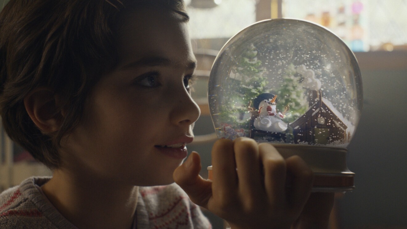 Regala alegría es la campaña de Navidad 2022 de Amazon. En la foto, un primer plano de la cara de una niña con el pelo coroto agarrando una bola de nieve de cristal. Dentro de la bola hay dos árboles negros, un muñeco de nieve sentado en un banco y una casa. Todo el paisaje está cubierto de nieve y también está nevando. 