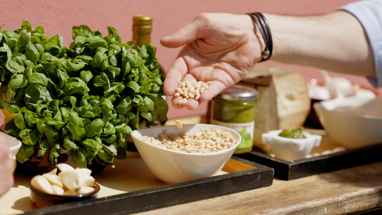 Es un bodegón de los ingredientes de la salsa al pesto: aceite, ajo, albahaca y piñones. Una mano agarra los piñones de un cuenco blanco y los deja caer. 