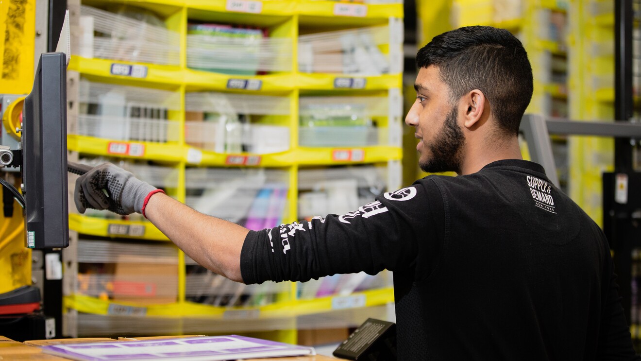 A man at an Amazon fulfillment center touches a monitor at his work station