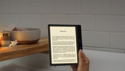 A woman holds a Kindle Oasis device with the page of a book on the display. Her arm appears from bubbles in a tub, with a wooden tub caddy behind the device.