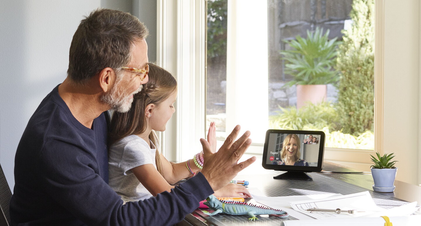 A little girl sits on a man's lap at a desk. They are both waving and looking at an Echo Show device, on which a woman is smiling and engaging with them. 
