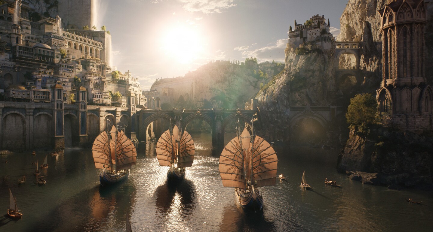 A scene from the Lord of the Rings: The Rings of Power. Ships with circular sails move through a bay. There is a bridge and a city on the hill surrounding the bay.