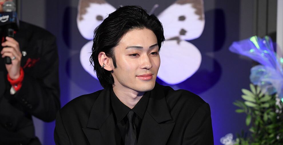 大きな蝶のモチーフや青い花が設置された青い壁を背に、微笑む話す男性。黒いスーツ、黒いシャツ、黒いネクタイを着用。
