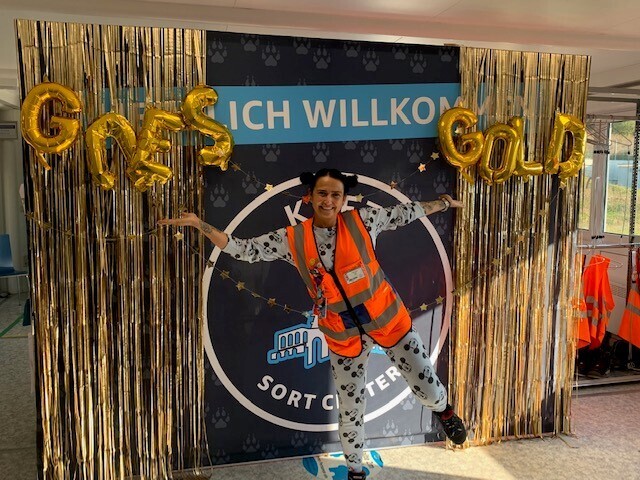 Eine Mitarbeiterin steht im Schlafanzug im Sortierzentrum in Wolfhagen vor einem Bild auf dem "Amazon goes gold" steht.