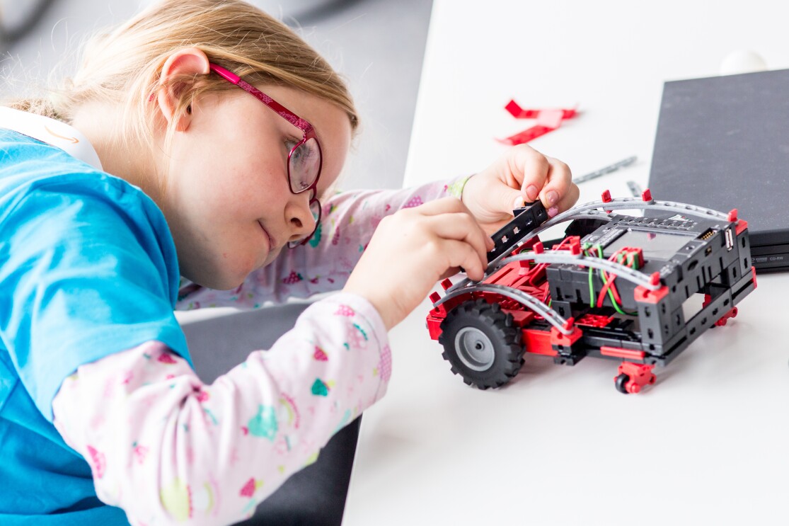 Ein kleines Mädchen mit Brille blickt auf einen mit Fischertechnik gebastelten Mini-Roboter, der auf einem Tisch steht