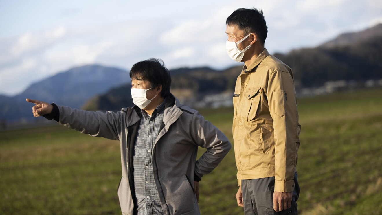 水田を背に画面左側の先を指さす木下さんと同じ方向を向いている松井さん