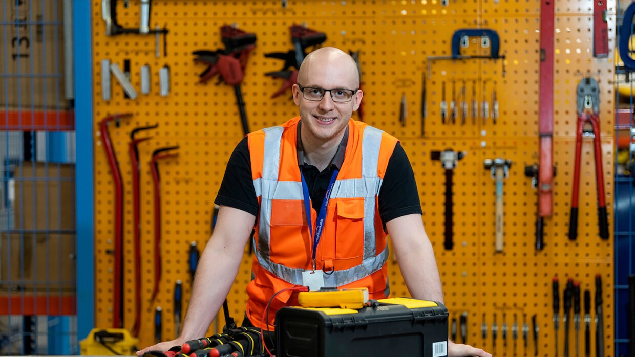 Luke Vials standing with his toolbox at his workbench wearing an orange high-viz vest.