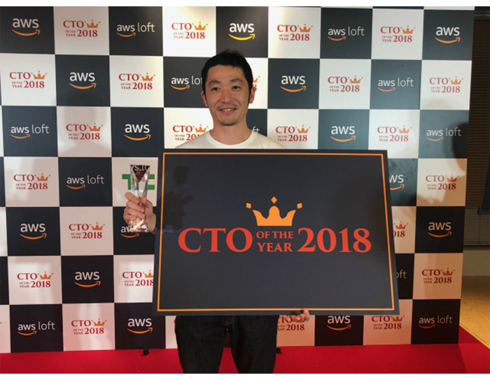 白と黒の市松模様の壁を背に立つ男性。手に透明なトロフィーと黒いボードを持っている。