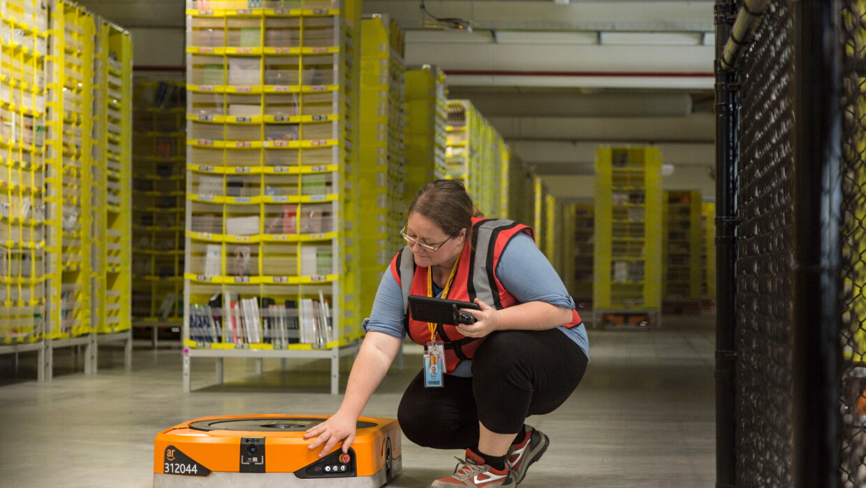 Foto di Maria Stella Carnà che regge un tablet di controllo e si china su uno dei robot di cui è responsabile all'interno del Centro di Distribuzione Amazon di Passo Corese