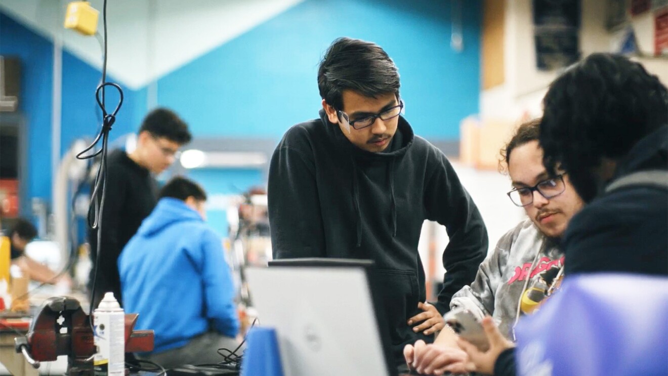 A photo of three students reviewing information on a laptop in a high school robotics lab.