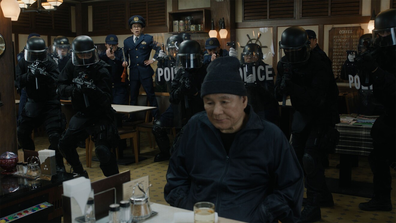 古びた喫茶店の中で、ニット帽をかぶって座っている黒づくめの男(ビートたけし)、その周囲を機動隊がライフルや盾を持って囲んでいる