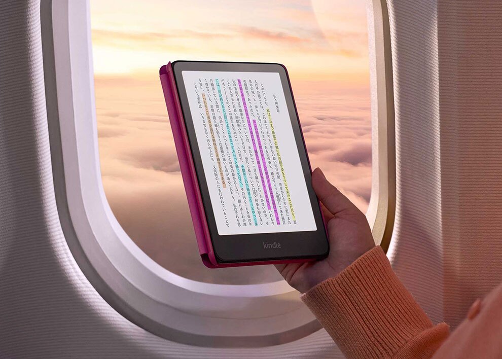 飛行機の窓際で、黒いフレームのKindle端末を片手に持っている。夕焼けの雲海が見える窓を背景に、画面には複数色のハイライトが入った縦書きの日本語ページが表示されている。