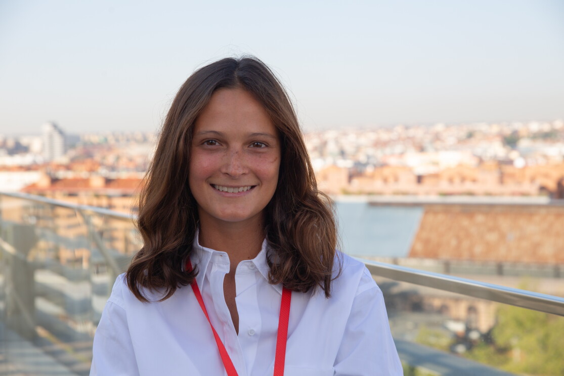 María Robert, Senior Program Manager de Experiencia del Cliente. De fondo el paisaje de Madrid. Está en el último piso de las oficinas corporativas. Hay una barandilla de color metal. María tiene el pelo castaño un poco más allá del hombro. Lo lleva suelto. Está morena, tiene pecas y lleva una camisa blanca. 