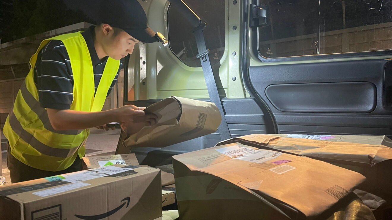 車の中で安全ベストを着ている男性が商品が入った紙袋を確認している。その紙袋を帽子に付けられたライトが照らしている