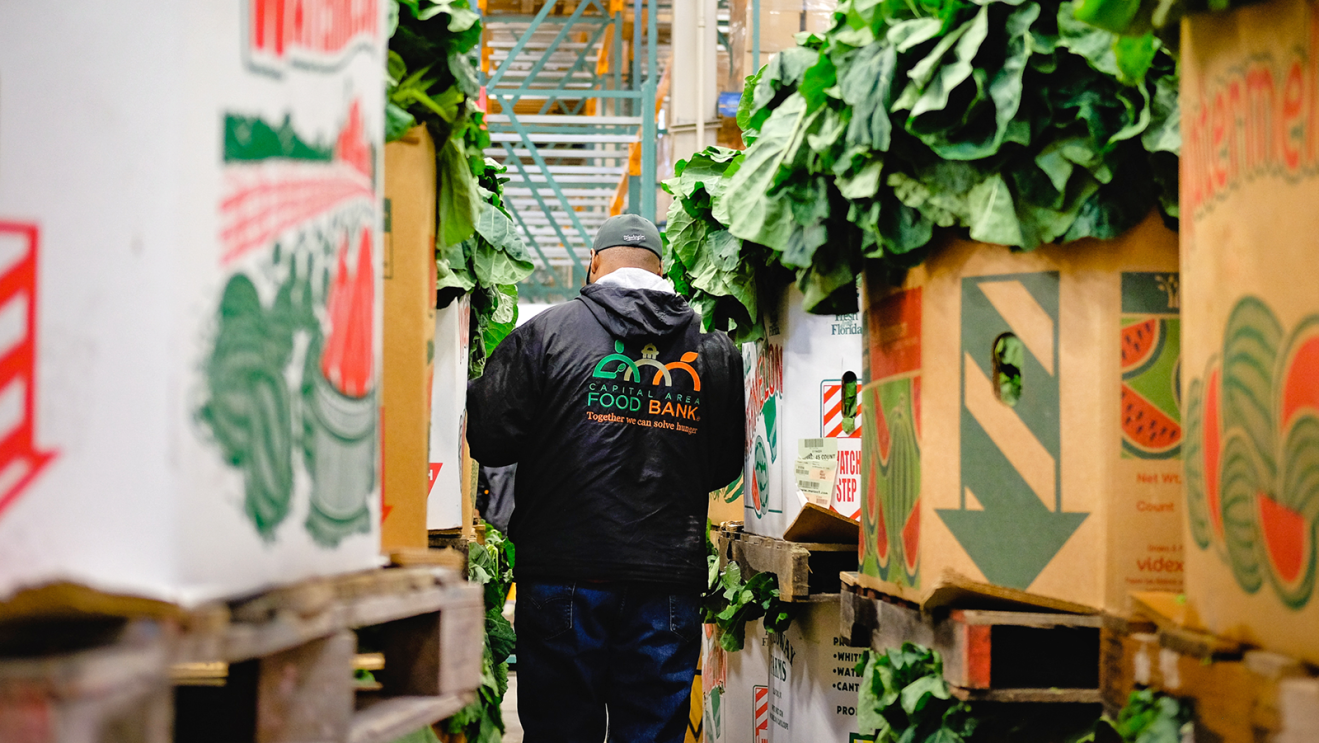 The back of a Food Bank worker in a warehouse. There are boxes of lettuce on either side of them.