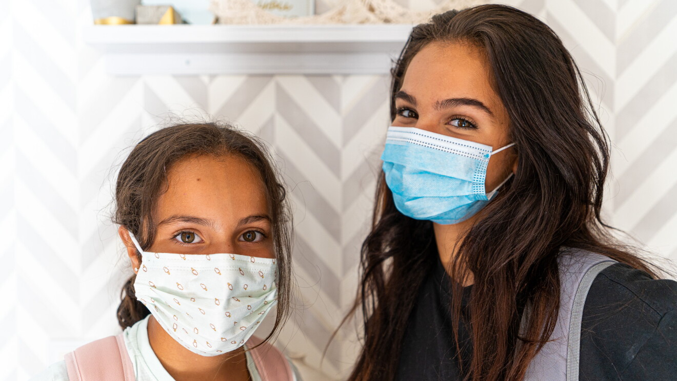Manuela y Martina con mascarilla y preparadas para salir para el cole.  Manuela a la derecha con una camiseta negra, una mochila moranda y el pelo suelo. Martina con una camiseta blanca, una mochila de color rosa y una mascarilla de ropa. Las dos miran a cámara. 