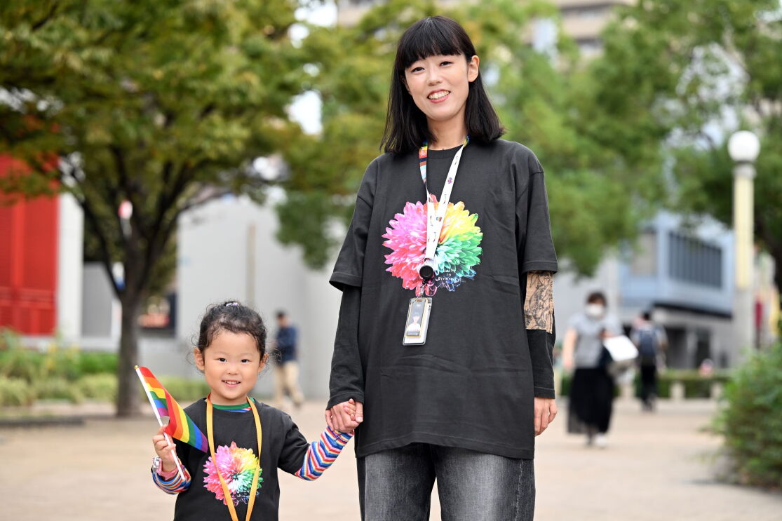 カラフルな花が描かれている黒いTシャツを着ている女性と女の子が公園で手を繋いで微笑んでいる。女の子は小さなレインボーフラッグを持っている。