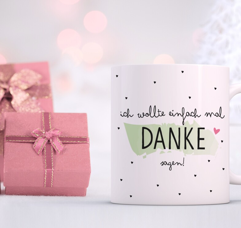 Eine weiße Tasse mit der Aufschrift "Ich wollte einfach mal Danke sagen" steht vor einem weißen Hintergrund mit rosafarben eingepackten Geschenken.