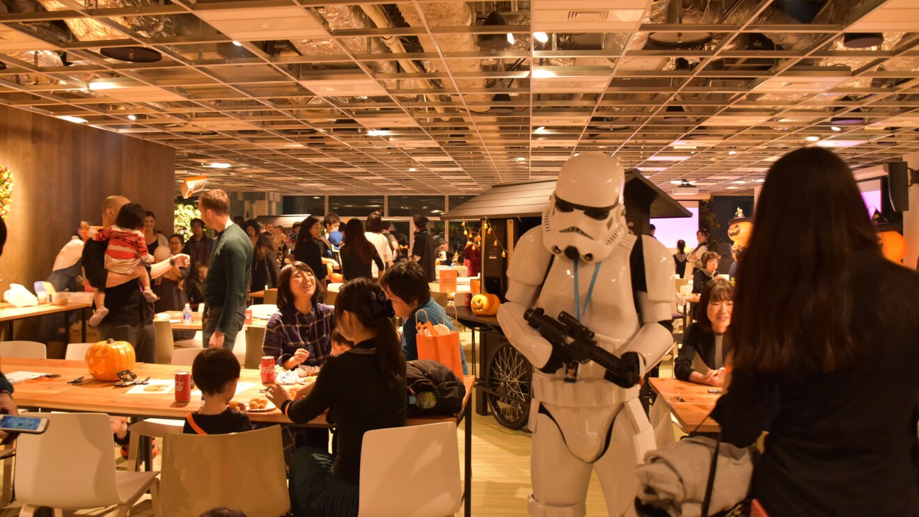 広いカフェスペースで食事や会話を楽しむ人々。中央の人物は白いアーマースーツを着用し、モデルガンを持っている。