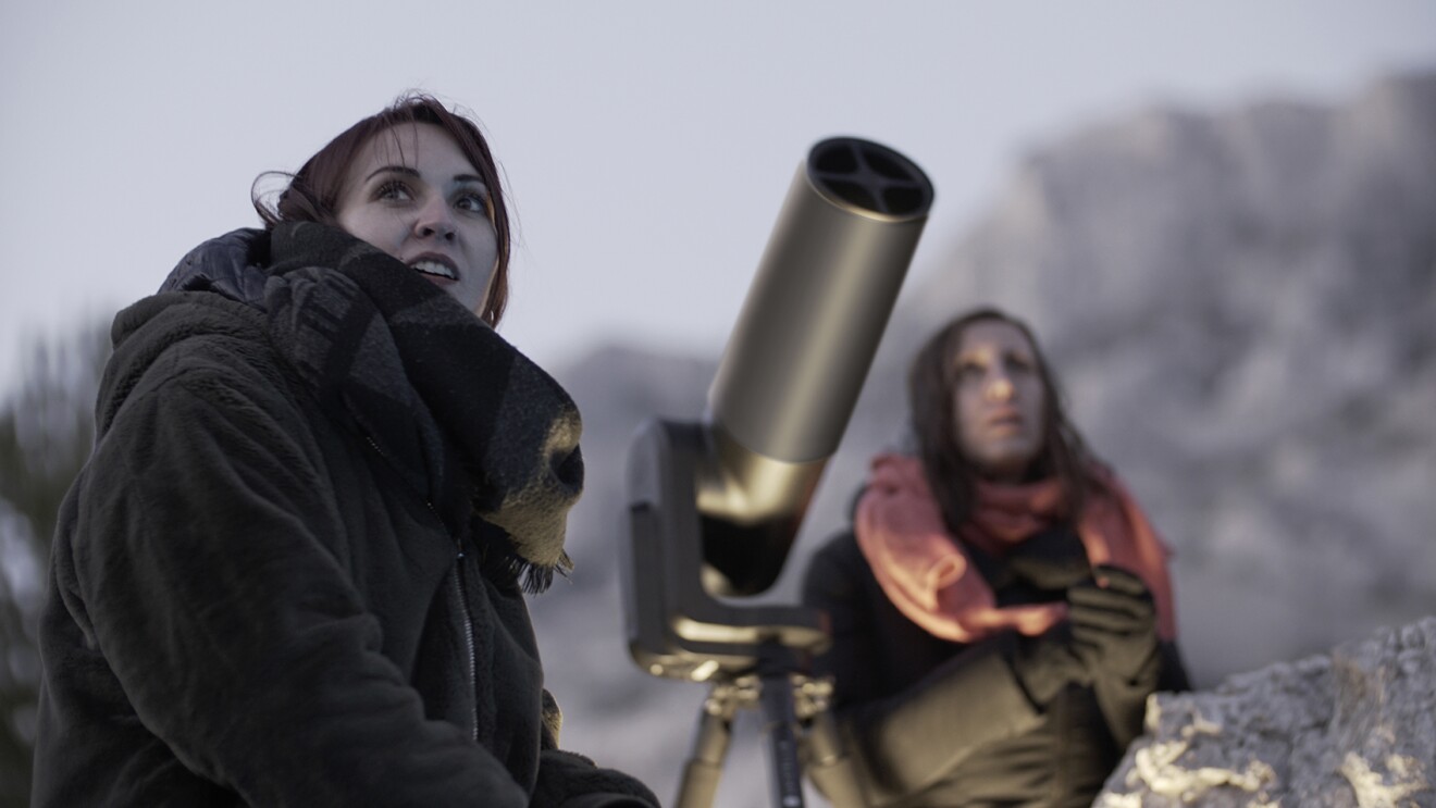 An image of two women sitting next to a telescope outside looking up at the sky above.