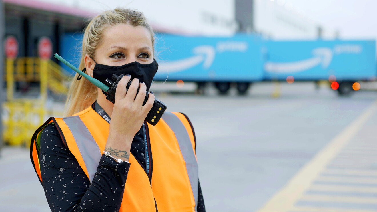 Ramona Moraru, supervisora de operaciones en la estación logística de Getafe. En el exterior del centro logístico con dos camiones de Amazon Prime de color azul con la sonrisa de Amazon en blanco. Ramona está hablando por un walkie talkie. Tiene el pelo claro, recogido en una cola, Con la mano derecha agarra el walkie talkie, tiene las uñas pintadas de negro. Lleva un tatuaje en la muñeca izquierda y un pulsera. Los ojos oscuros y maquillados y una mascarilla negra de Amazon. Lleva un chaleco de color naranja y un jersey negro con estrellas de color gris.  
