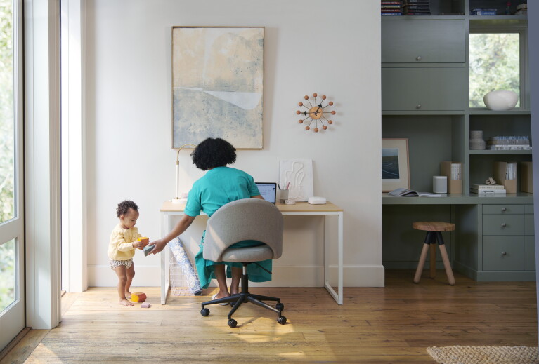 A woman sits at a desk, helping her toddler. On her desk is an Eero device.