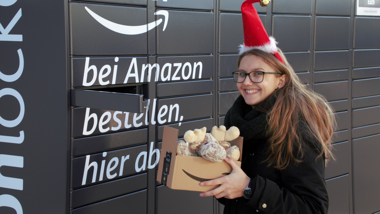 Eine Frau mit Brille und langen Haaren hält ein Amazon Karton vor einem Locker. Sie trägt eine rote Nikolausmütze. im Paket ist ein Elch zu sehen.
