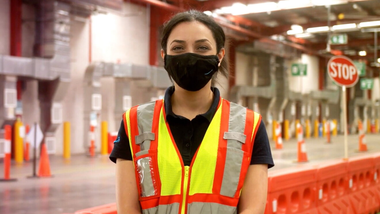 Daniella, an operations lead, stands in a fulfilment centre wearing a mask and a safety vest, looking at the camera.