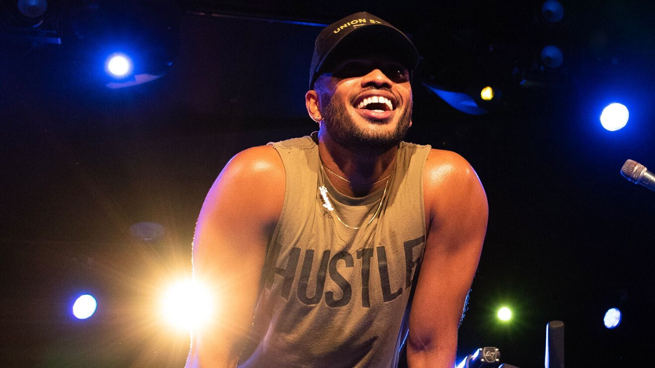 An image of a man smiling while looking out at a group of people. There is a stationary bike behind him and stage lighting.