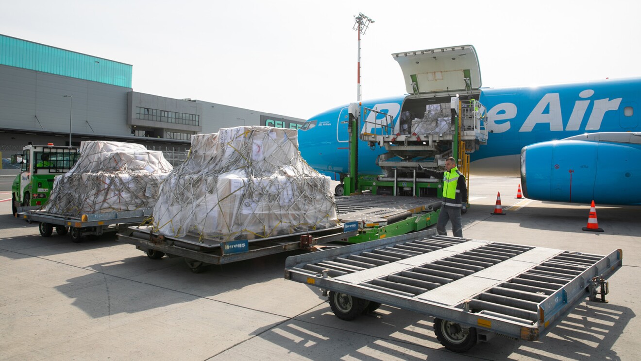 Pallets of humanitarian goods are loaded onto an Amazon Air cargo plane. A man is watching and giving a thumbs up, as if to give an all clear to allow for the pallet to move towards the plane.