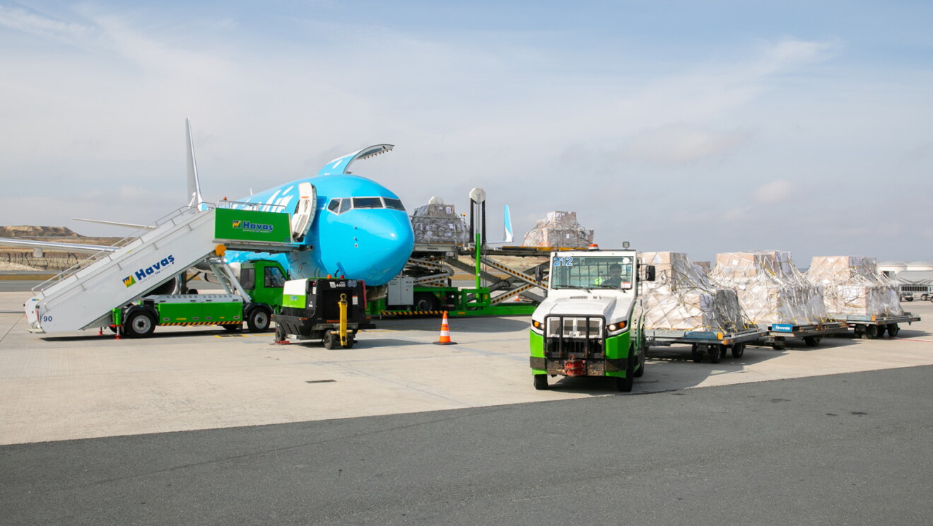 En la pista de un aeropuerto hay cuatro palés cargados con ayuda humanitaria. Los palés son de papel marrón, con pegatinas blancas y unas cuerdas. De fondo un avión de color azul con las letras en blanco Prime Aire. El cielo está nublado y es el atardecer. Los palés están arrastrados por una camioneta verde y blanca y el avión tiene una escalera por un lado y por el otro ujna grúa por la que se está cargando la ayuda humanitaria. 