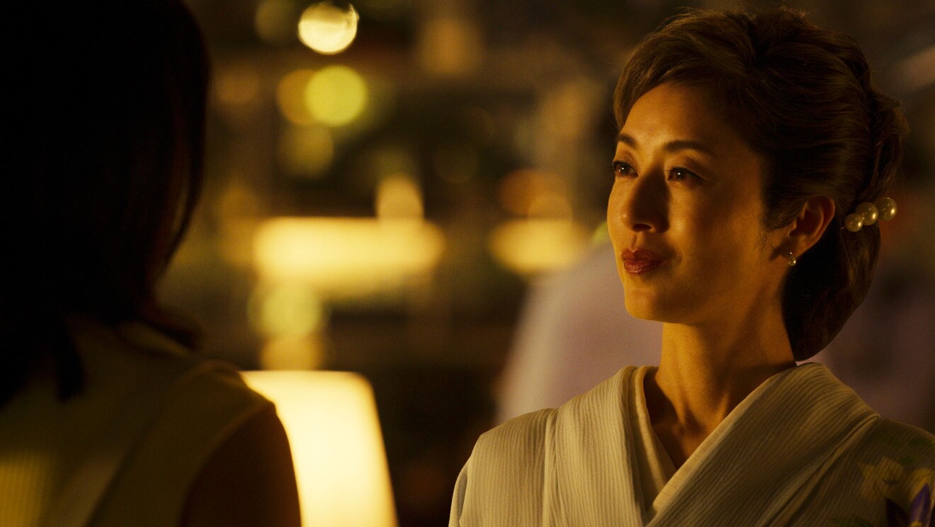 黄色いライトに照らされた室内にいる着物姿の女性が、対面に立つ女性を見つめいている