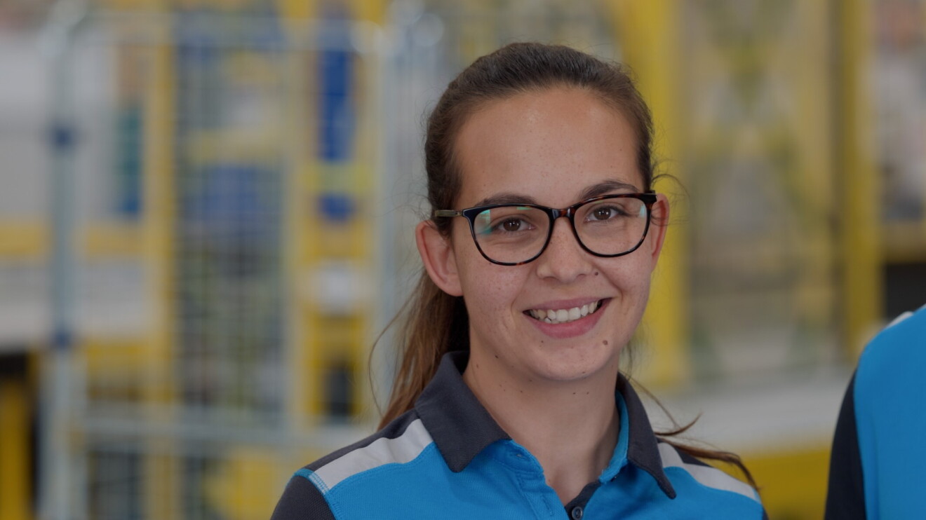 Maria de Dolinski - Becerra Services, S.L. mirando a cámara con una sonrisa. Lleva el pelo largo pero recogido en una coleta, unas gafas de pasta marrones, tiene pecas en la cara y lleva un jersey de colores azules. De fondo se ven estanterías de Amazon. 
