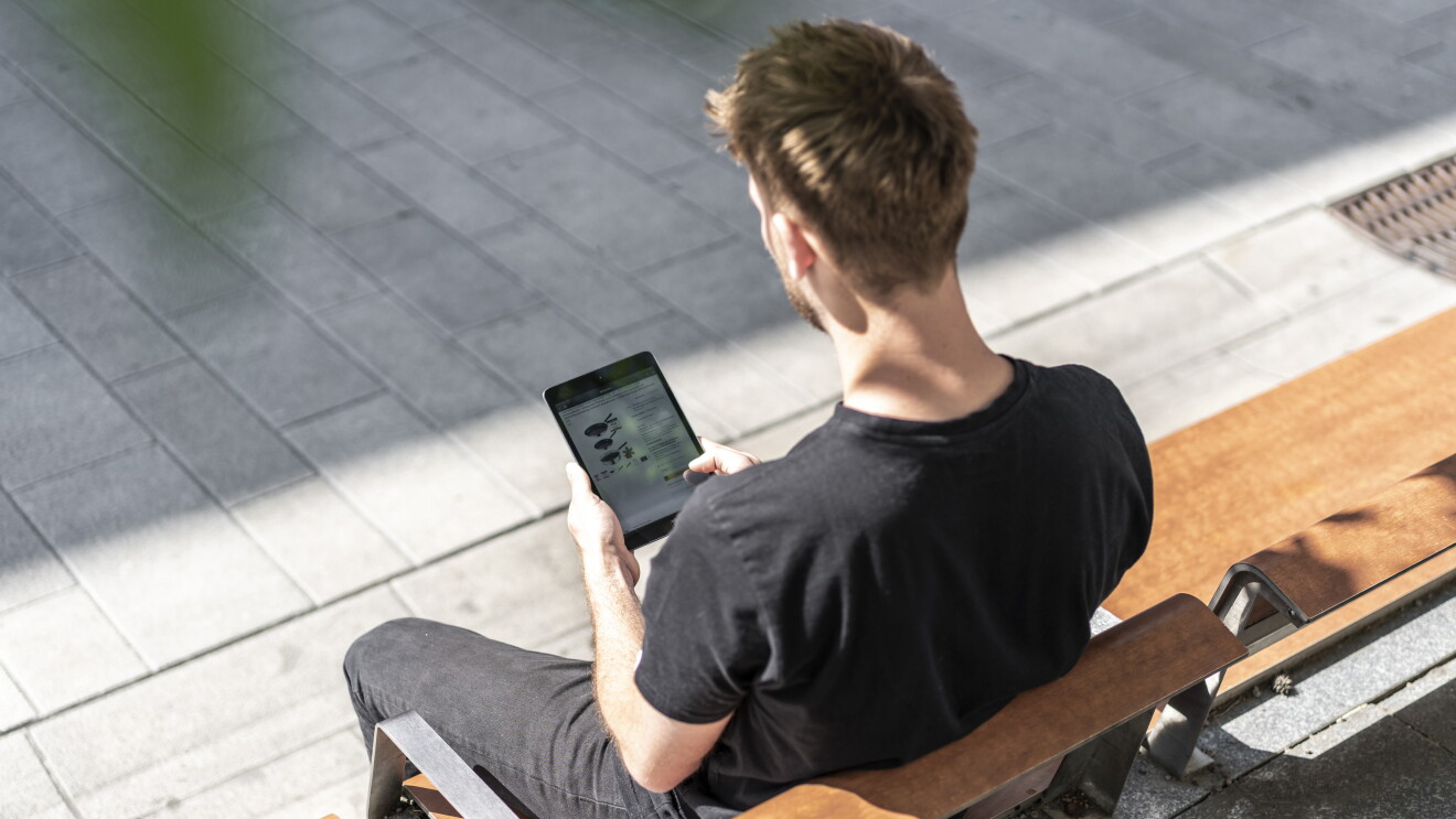 Ein junger Mann sitzt auf der Parkbank und hat ein Tablet in der Hand. Er bestellt von unterwegs bei Amazon.