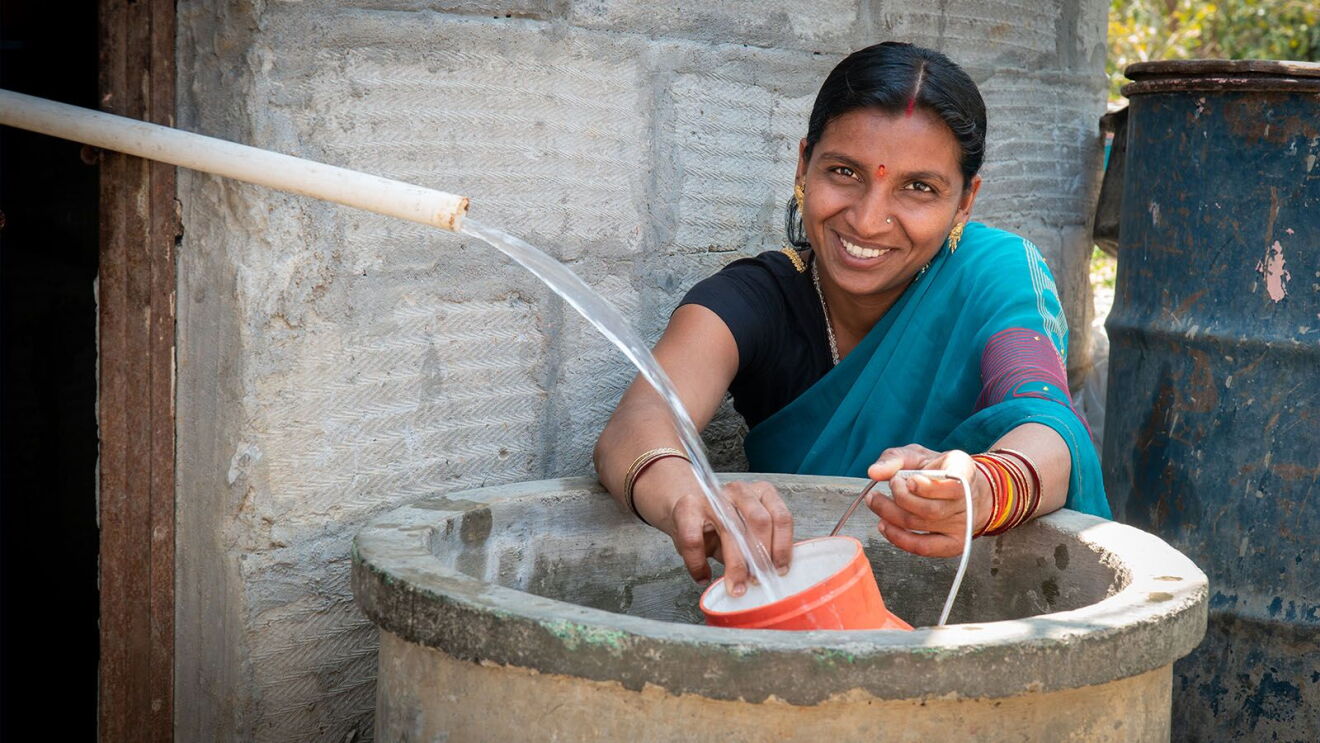 Woman in turquoise sari filling an orange container with water from a pipe at a concrete well