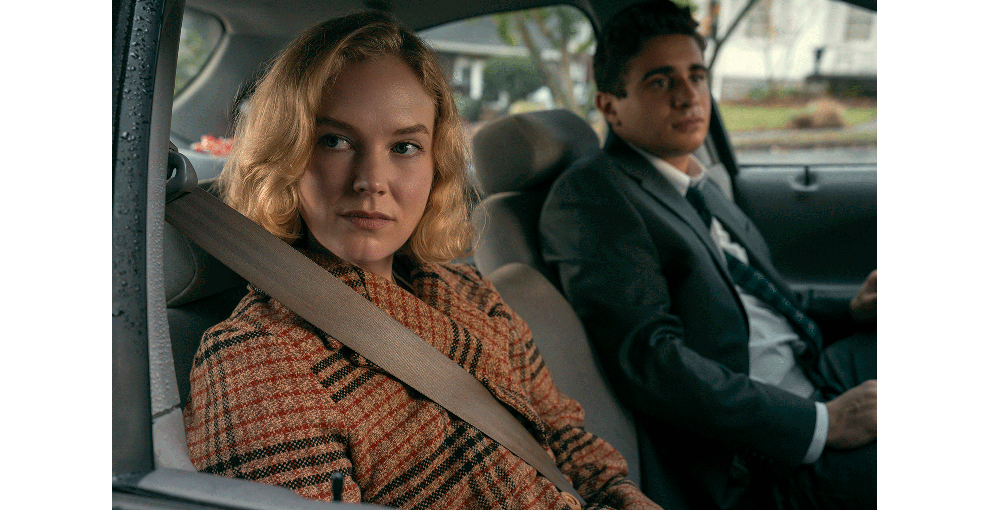 車の座席に並んで座る、チェック柄のコートを着た金髪の女性と、ダークスーツを着た黒髪の男性。女性だけシートベルトをしている。