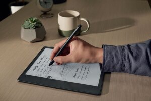 An image of a handing writing on a Kindle Scribe device using a stylo pen made for the device. There is a small plant and a coffee mug on the desk in the background.