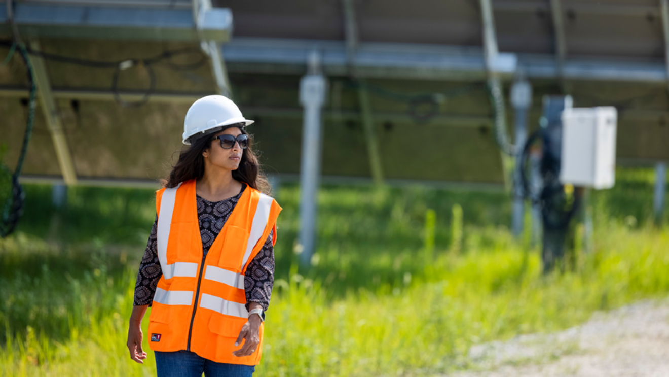 En una granja solar, delante de la parte trasera de tres placas aparece caminando una mujer con gafas de sol, casco blanco, y el pelo ondulado largo. Lleva una jeans, un chaleco de color naranja con un jersey negro estampados con formas geométricas grises.
