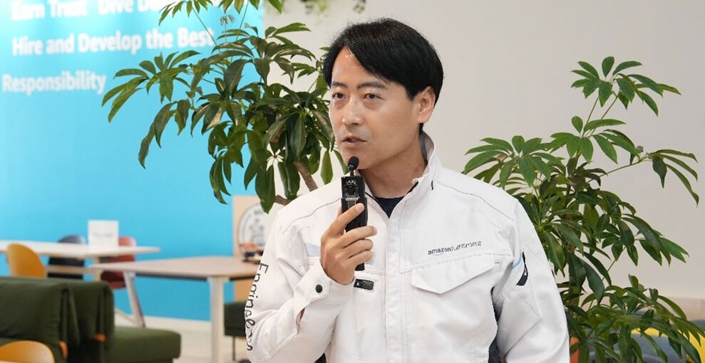 白い壁と観葉植物を背景に、白い作業着姿の人物がマイクを手に話している。奥の方の壁は水色で、テーブルや椅子が並んでいる。