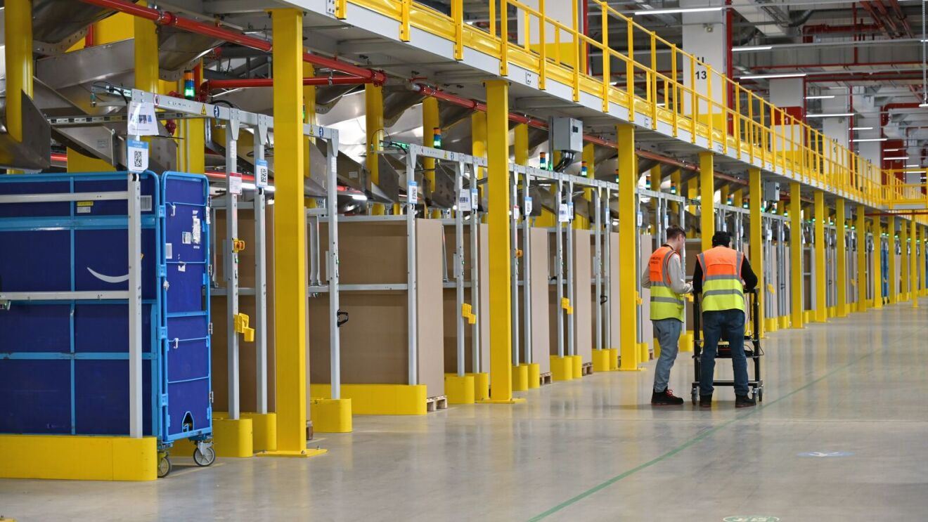 Zwei Logistikmitarbeiter mit orange-gelber Veste in einer Halle mit gelben Säulen.