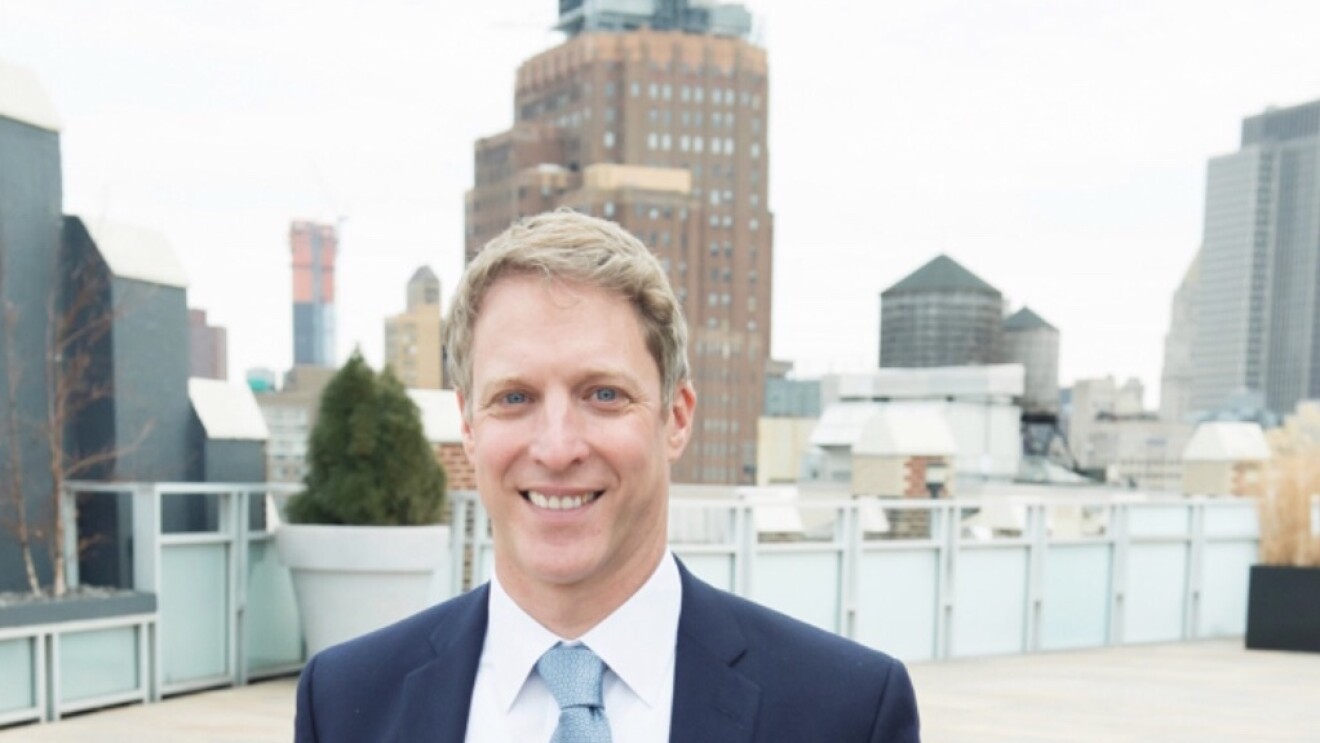 A man in a navy suit, white shirt, and slate blue tie stands outdoors in a city setting.